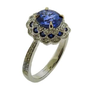 18 karat white gold ring with a beautiful vintage scalloped halo design. The ring featuring milgrain details for an addition vintage feel. It is set with a stunning sapphire and is accented with sapphires and diamonds in the halo.