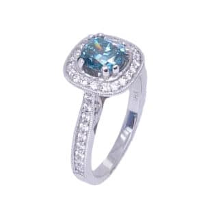 In 18 karat white gold, this vintage inspired halo ring showcases a 1.03 carat cushion cut treated blue diamond. Accented with round brilliant cut diamonds totaling 0.45 carats. The perfect gift for 10th anniversaries, 60th anniversaries, April birthdays or a stunning engagement ring!