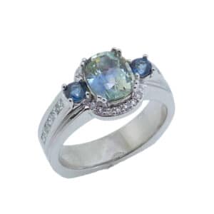 14K white gold custom halo ring by Studio Tzela set with a 1.48ct bi-colour sapphire and accented with 2 round blue/green sapphires, 0.19cttw, 12 = 0.273cttw G/H, VS-SI princess cut diamonds and 16 = 0.085cttw G/H, VS-SI round brilliant cut diamonds in the halo. This stunning ring is a custom design by David.  Sapphire is the birthstone for September.