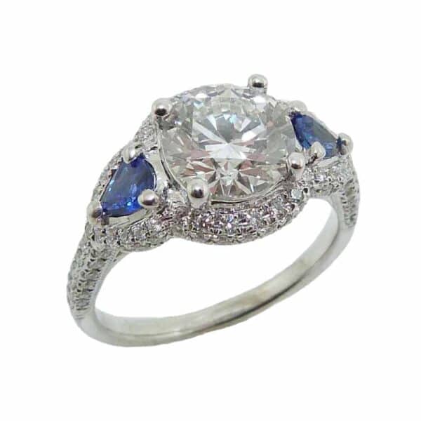 14K White gold diamond and sapphire engagement ring claw set in the center with one 1.57 carat, D, VVS2 round brilliant lab grown diamond. Claw set on the sides with two pear shaped blue sapphires totaling 0.39 carats and accented with 104 round brilliant cut diamonds totaling 0.40 carats, SI, G/H. Available in 14K gold, 18K gold, or platinum. This ring can be made in any combination of white, pink or yellow gold and can be customized to accommodate different size and shape diamonds, by special order.