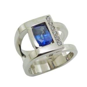 14K White gold custom lady's ring by Studio Tzela bezel set in the centre with a 1.55 carat elongated cushion blue sapphire and accented on the side and profiles with ten very good cut, natural round brilliant cut diamonds, 0.113 total carat weight, G/H, SI.