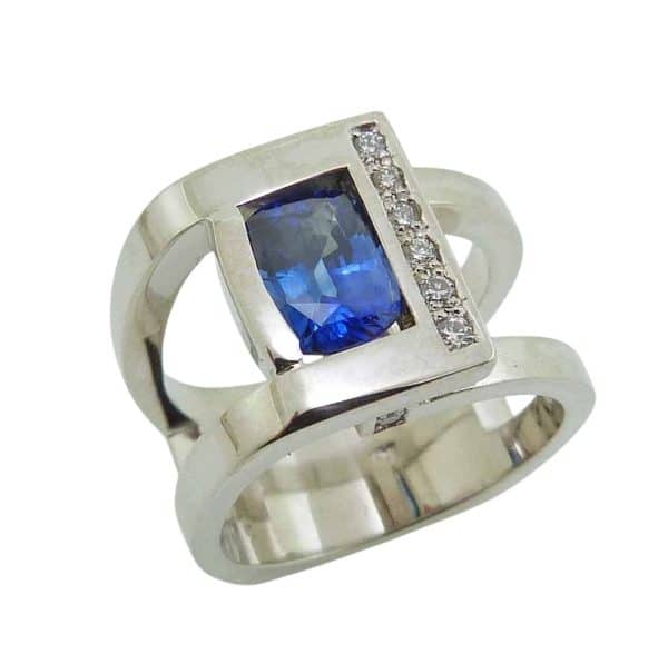 14K White gold custom lady's ring by Studio Tzela bezel set in the centre with a 1.55 carat elongated cushion blue sapphire and accented on the side and profiles with ten very good cut, natural round brilliant cut diamonds, 0.113 total carat weight, G/H, SI.